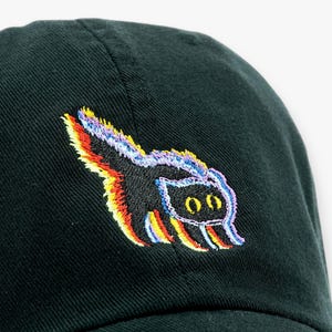 May include: A dark green baseball cap with a colorful embroidered skunk design. The skunk has a black body with rainbow-colored accents and two oval eyes.