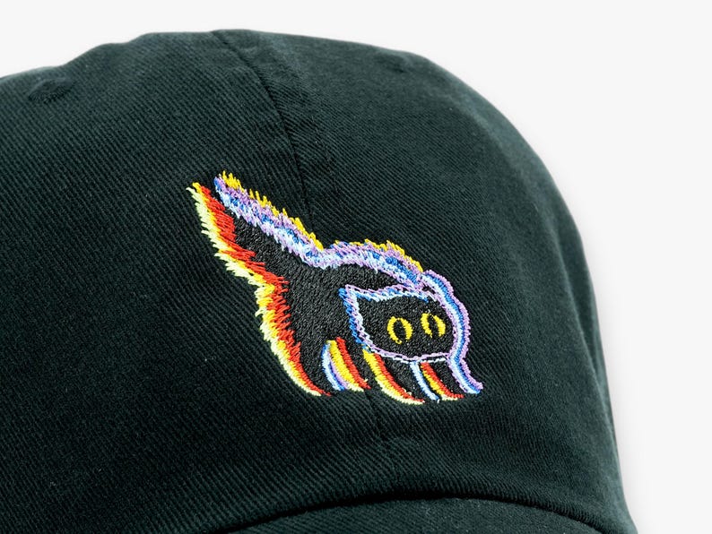May include: A dark green baseball cap with a colorful embroidered skunk design. The skunk has a black body with rainbow-colored accents and two oval eyes.