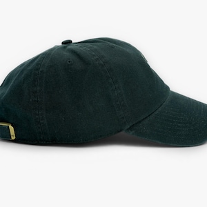 May include: A dark green baseball cap with a curved brim and adjustable strap. The cap has a metal buckle and a small button on top. The hat is made of a soft, textured fabric.