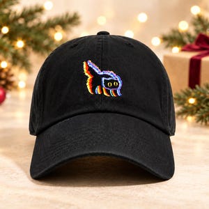 May include: A black baseball cap featuring an embroidered graphic of a cat with a glitch effect. The cat design incorporates vibrant rainbow colors including red, orange, yellow, blue, and purple, set against the solid black fabric of the cap.
