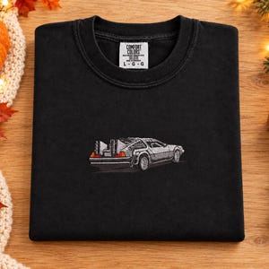 Puede incluir: Una camiseta negra de cuello redondo con un coche deportivo plateado bordado en la parte delantera. La etiqueta interior del cuello muestra el texto Comfort Colors, 100% Cotton, Made in Honduras y la talla L-G-G. La camiseta está doblada sobre madera.