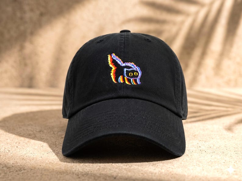 May include: A black baseball cap featuring an embroidered graphic of a cat with a glitch effect. The cat design displays vibrant, layered colors including red, blue, and yellow, with two large, round, yellow eyes centered on the front panel.