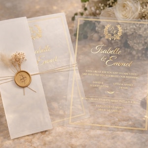 以下が含まれることがあります： 金色の文字が施されたエレガントな結婚式の招待状。「Isabelle & Emmet」の名前が記され、ワックスシールとドライフラワーで飾られています。結婚指輪と小さなトレイも写っています。