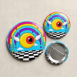 May include: Two circular pin-back buttons featuring a graphic of a yellow winged eyeball centered under a rainbow with dripping paint effects. The background shows a blue sky with white clouds above a black and white checkered floor pattern.