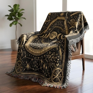 May include: A black and gold woven throw blanket featuring a celestial design with a central moon phase, stars, constellations, and a lion figure. The rectangular blanket has a decorative fringe along the edges and is draped over a chair.