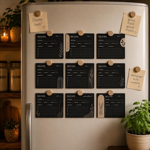 Könnte beinhalten: Neun schwarze, rechteckige Rezeptkarten mit weißem Text und Liniengrafiken, befestigt mit runden Holzmagneten an einem Kühlschrank. Drei beige Haftnotizen mit handschriftlichem Text sind ebenfalls angebracht: 'Dinner tues!', 'good food good mood' und 'don't forget to be awesome'.