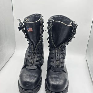 Può includere: Un paio di stivali tattici in pelle nera con chiusura a zip frontale, lacci neri e punta rinforzata. Una piccola toppa con la bandiera americana e la scritta MADE IN USA è visibile sulla linguetta interna dello stivale sinistro.