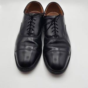 May include: A pair of black leather oxford shoes featuring a cap toe design, thin black laces, and a tan interior lining. The shoes have a smooth, polished finish with visible stitching along the cap toe and eyelet panels, set on a dark sole.