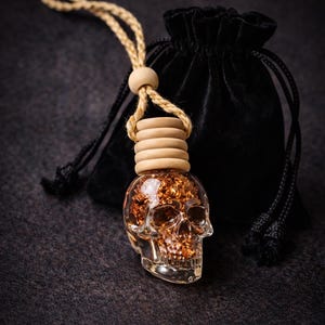 Puede incluir: Una botella transparente en forma de calavera llena de purpurina dorada, coronada con anillos de madera y una cuerda beige. Una bolsa de terciopelo negro con cordón está en el fondo. El objeto es decorativo.
