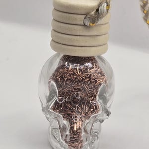 May include: A clear glass skull-shaped bottle filled with copper-colored material, topped with a light wood cap and a braided rope. The bottle has a decorative design and is likely an air freshener or diffuser.