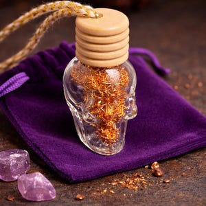 Puede incluir: Una botella de vidrio transparente con forma de calavera llena de alambre de color cobre, coronada con una tapa de madera. La botella está sobre una bolsa de terciopelo morado, con dos cristales rosas cerca. Un cordón dorado y blanco está unido.