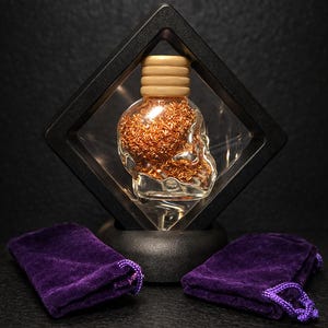 May include: A glass skull-shaped bottle filled with copper-coloured metal pieces, presented in a black shadow box. The bottle has a wooden cap. Two small, purple velvet drawstring bags are in front of the display.