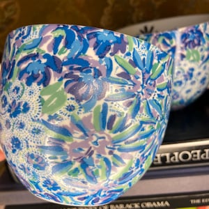 May include: Close-up of a ceramic cup with a floral pattern. The cup features a white base with a design of blue, light blue, and purple flowers and green leaves. The cup is sitting on a stack of books.