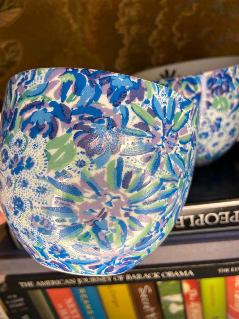 May include: Close-up of a ceramic cup with a floral pattern. The cup features a white base with a design of blue, light blue, and purple flowers and green leaves. The cup is sitting on a stack of books.