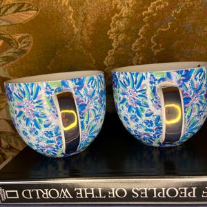 May include: Two ceramic mugs with a blue, green, and purple floral pattern. The mugs have a silver handle and a white interior. The mugs are sitting on top of a stack of books.