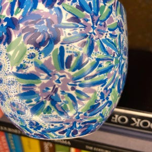 May include: A ceramic tumbler with a floral pattern in shades of blue, purple, and green. The design features stylized flowers and leaves against a white background. The tumbler is cylindrical and appears to be sitting on a shelf.