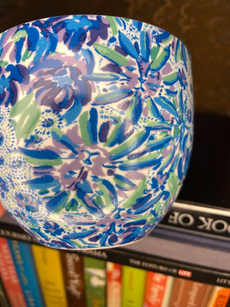 May include: A ceramic tumbler with a floral pattern in shades of blue, purple, and green. The design features stylized flowers and leaves against a white background. The tumbler is cylindrical and appears to be sitting on a shelf.