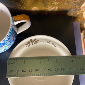 May include: A white saucer with a gold floral design and the text "xx. Lilly". The saucer is approximately 4 inches in diameter. A blue and white floral teacup with a brown handle is partially visible in the background.