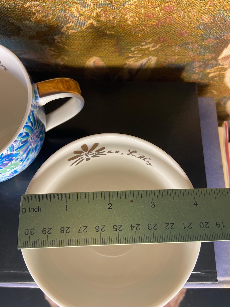 May include: A white saucer with a gold floral design and the text "xx. Lilly". The saucer is approximately 4 inches in diameter. A blue and white floral teacup with a brown handle is partially visible in the background.