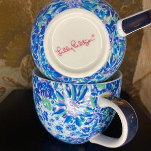 May include: Two stacked ceramic mugs with a vibrant floral pattern in shades of blue, green, and purple. The mugs have a white interior and a dark blue handle. The bottom mug is visible, and the brand name "Lilly Pulitzer" is printed on the bottom of the top mug.