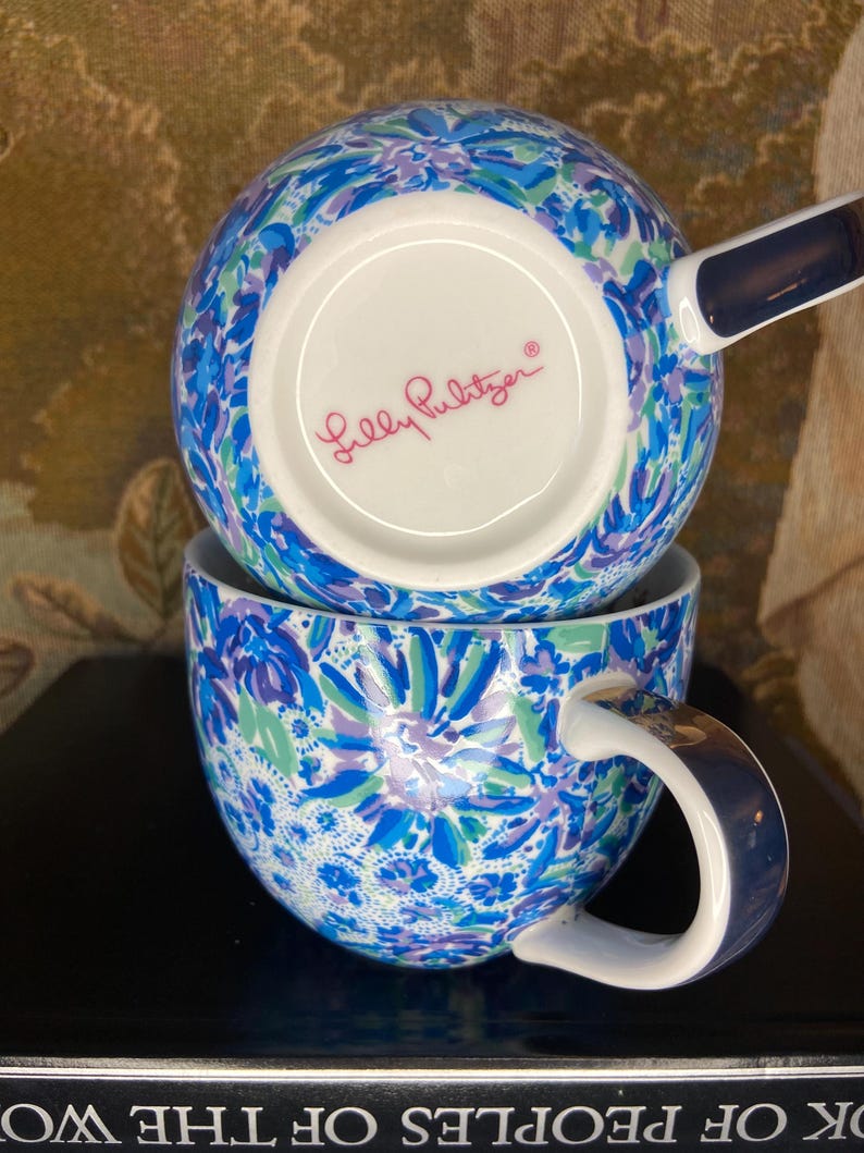 May include: Two stacked ceramic mugs with a vibrant floral pattern in shades of blue, green, and purple. The mugs have a white interior and a dark blue handle. The bottom mug is visible, and the brand name "Lilly Pulitzer" is printed on the bottom of the top mug.