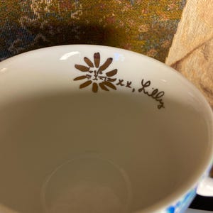 May include: White ceramic mug with a floral design in gold and the text "xx, Lilly". The bottom of the mug features a blue and purple floral pattern. The mug is designed for beverages.