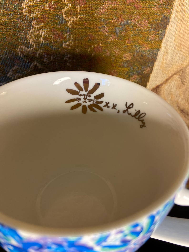 May include: White ceramic mug with a floral design in gold and the text "xx, Lilly". The bottom of the mug features a blue and purple floral pattern. The mug is designed for beverages.