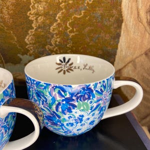 May include: A pair of ceramic mugs with a vibrant floral pattern in shades of blue, purple, and green. The mugs have white handles and interiors. The interior of one mug has a gold floral design and the text "xx, Lilly".