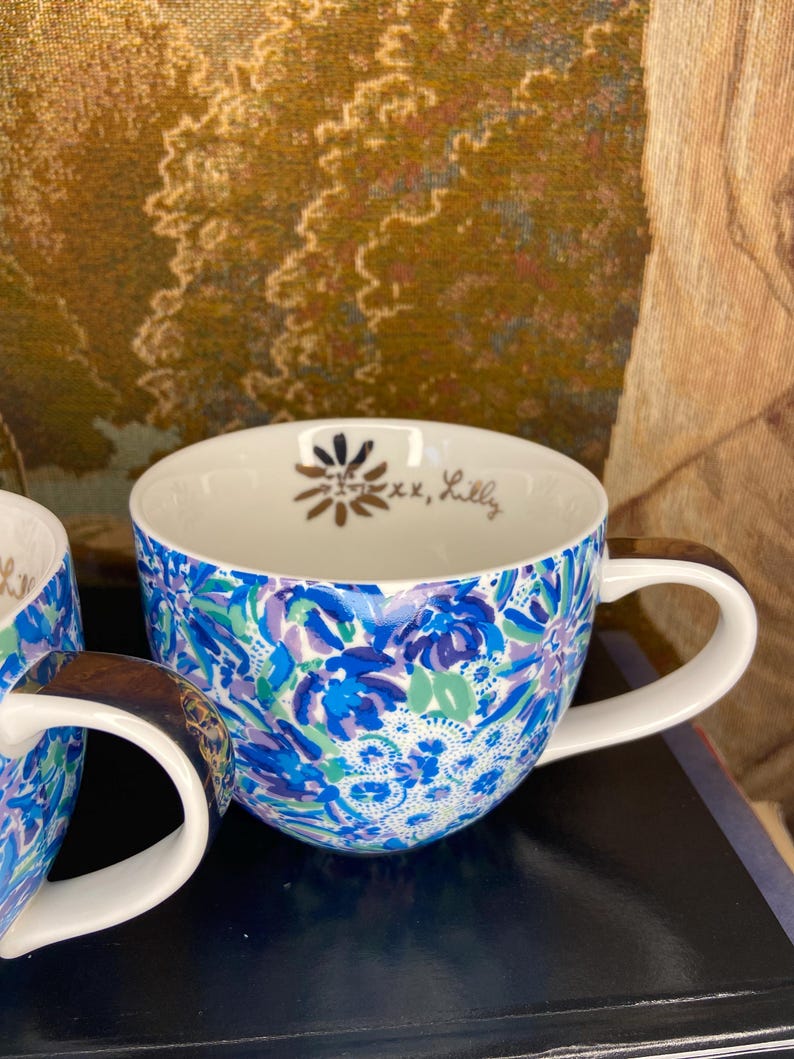 May include: A pair of ceramic mugs with a vibrant floral pattern in shades of blue, purple, and green. The mugs have white handles and interiors. The interior of one mug has a gold floral design and the text "xx, Lilly".