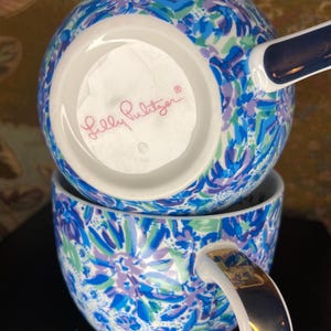 May include: Two stacked ceramic mugs with a floral pattern in shades of blue, green, and purple. The mugs have white interiors and handles with gold accents. The bottom mug's handle is visible, and the bottom of the top mug shows the "Lilly Pulitzer" logo.