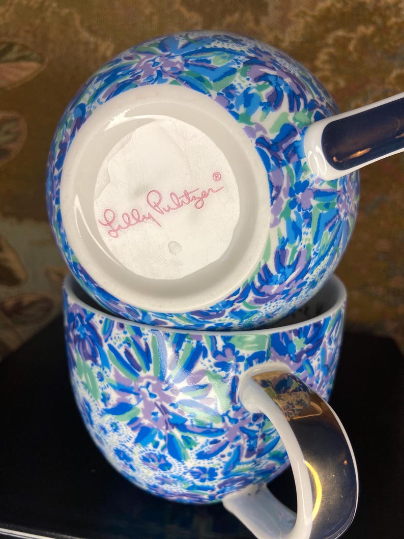 May include: Two stacked ceramic mugs with a floral pattern in shades of blue, green, and purple. The mugs have white interiors and handles with gold accents. The bottom mug's handle is visible, and the bottom of the top mug shows the "Lilly Pulitzer" logo.