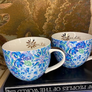 May include: Two white-handled ceramic mugs with a blue floral pattern. The mugs have a gold floral design and the text "xx, Lilly" inside. The mugs are sitting on top of a stack of books.
