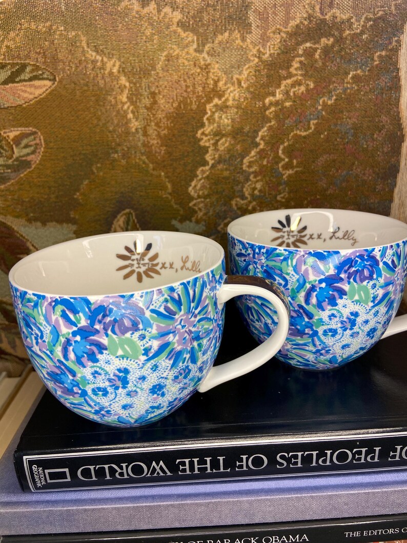 May include: Two white-handled ceramic mugs with a blue floral pattern. The mugs have a gold floral design and the text "xx, Lilly" inside. The mugs are sitting on top of a stack of books.
