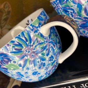 May include: Two ceramic mugs with a floral pattern in shades of blue, green, and purple. The mugs have white handles and a glossy finish. The design features stylized flowers and leaves. The mugs are likely for beverages.