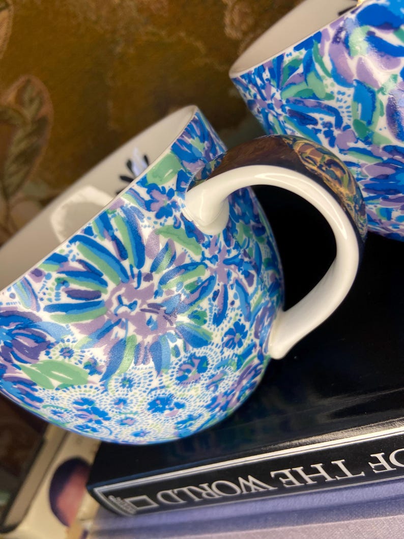 May include: Two ceramic mugs with a floral pattern in shades of blue, green, and purple. The mugs have white handles and a glossy finish. The design features stylized flowers and leaves. The mugs are likely for beverages.