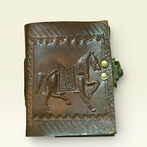 May include: A brown leather journal with a detailed embossed image of a horse. The journal has a decorative border and a metal clasp closure with two brass-colored rivets. The leather has a distressed appearance.