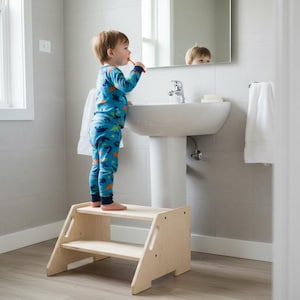 May include: A child in blue dinosaur pyjamas brushes their teeth at a bathroom sink, standing on a light wood step stool. The bathroom has a white sink, mirror, and towel rail. The floor is a light wood colour.