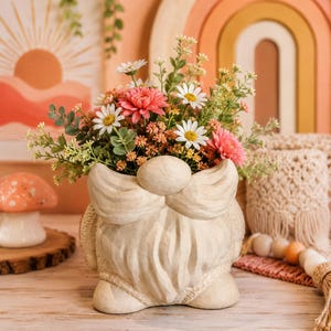 May include: A whimsical gnome-shaped planter filled with a vibrant arrangement of pink, white, and green flowers. The off-white planter has a textured finish and is decorated with a bow and beard. The background includes a wooden rainbow and a sun-themed artwork.
