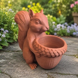 Könnte beinhalten: Ein rotbrauner Eichhörnchen-Blumentopf aus Terrakotta, der eine eichelförmige Schale hält. Das Eichhörnchen steht auf einem Steinweg, umgeben von Blumen und Grün. Der Topf ist für den Außenbereich geeignet.