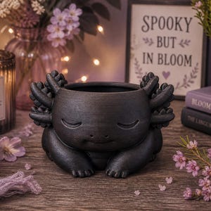 May include: A dark gray axolotl-shaped planter with closed eyes, featuring detailed gills and feet. The planter is sitting on a wooden surface, with a candle, flowers, and a sign that reads "Spooky But In Bloom" in the background.