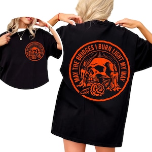 Puede incluir: Camiseta negra con un diseño circular naranja en la parte delantera y trasera. El diseño presenta una calavera, velas y una rosa, con el texto "MAY THE BRIDGES I BURN LIGHT MY WAY" rodeando el diseño. Camiseta de estilo casual.