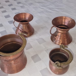Può includere: Una collezione di quattro recipienti in rame che include due piccole brocche con bordi svasati e due secchi rotondi con manici oscillanti in ottone. Le superfici metalliche presentano una finitura calda con texture martellate visibili.