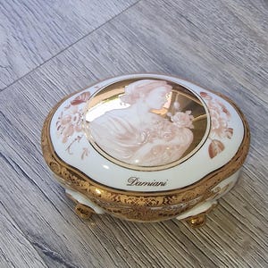 Może przedstawiać: Owalne pudełko na drobiazgi z porcelany z białą podstawą i złotym wykończeniem. Wieczko posiada centralną ilustrację w stylu kamei przedstawiającą osobę z profilu trzymającą kwiaty, otoczoną motywami roślinnymi. Słowo Damiani jest nadrukowane na wieczku.