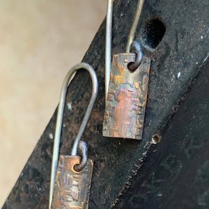 以下が含まれることがあります： ハンマーで叩かれた銅の四角いペンダントがついた、銀製のドロップイヤリングのペア。銅は、茶色、金色、赤色の色合いの、素朴な酸化仕上げになっています。