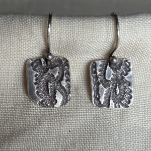 May include: A pair of silver-toned earrings. Each earring features a rectangular pendant with a textured, organic design. The earrings have a simple hook closure. The background is a light-colored fabric.