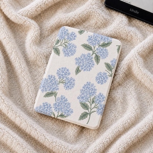 Könnte beinhalten: Eine rechteckige E-Reader-Hülle mit cremefarbenem Hintergrund und einem sich wiederholenden Muster aus hellblauen Hortensienblüten und grünen Blättern. Die Hülle hat abgerundete Ecken und eine glatte, matte Oberfläche mit Blumendesign.
