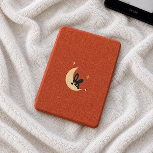 Peut inclure: Une housse de liseuse rectangulaire avec une finition en tissu orange brûlé texturé. Le centre présente un motif brodé d'une silhouette de lapin noir nichée dans un croissant de lune jaune pâle, accentué par de petites étoiles jaunes.