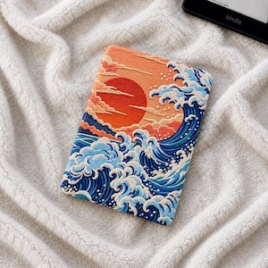 Peut inclure: Une housse de liseuse rectangulaire présentant une broderie d'un grand soleil rouge se levant sur des vagues océaniques bleues stylisées avec des crêtes d'écume blanche, sur un fond de nuages orange et pêche avec une finition en tissu texturé.