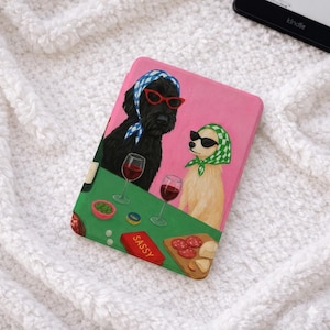 Peut inclure: Étui Kindle rose avec une illustration fantaisiste de deux chiens portant des lunettes de soleil et des foulards, dégustant du vin et des collations. Le mot "SASSY" est imprimé sur une boîte rouge. Un lecteur Kindle est visible en arrière-plan.