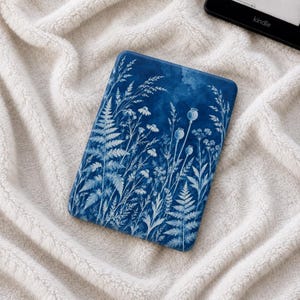 Peut inclure: Une housse de liseuse rectangulaire présentant un fond bleu profond avec un imprimé botanique blanc composé de fougères, de fleurs sauvages et de capsules de pavot. Le motif recouvre toute la surface avant de l'appareil posé sur un tissu blanc texturé.
